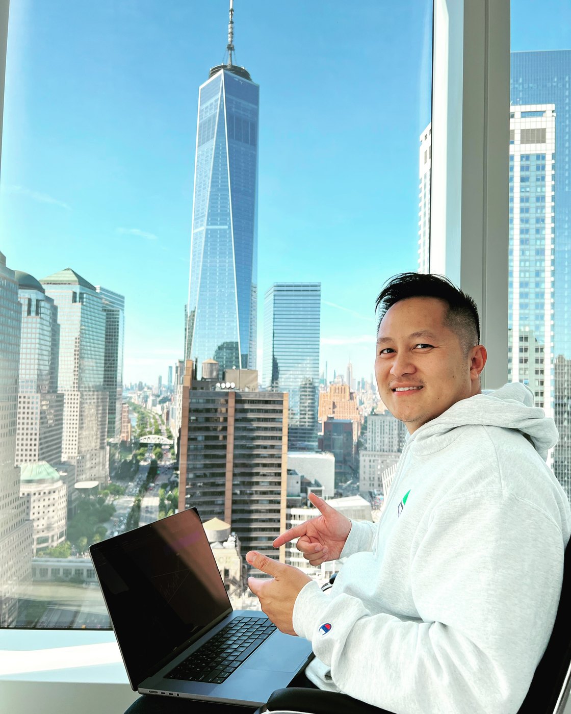 Tim Luong in a Champion hoodie with a laptop showing trading charts and a city skyline behind him